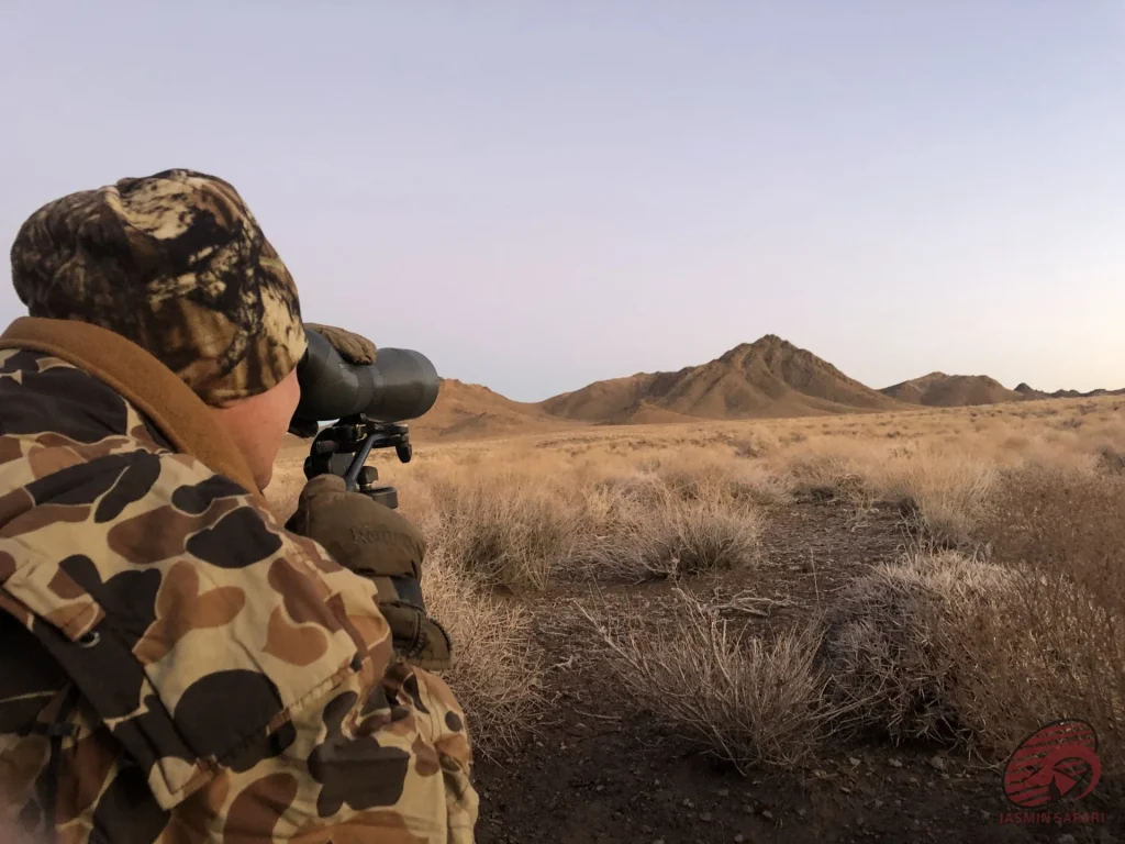 Hunter using a spotting scope at dawn on an Iranian desert plain with low mountains, hunt in Iran