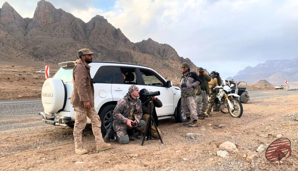 Hunters glassing with a spotting scope beside a white Suzuki 4×4 and motorcycle on a mountain road in Iran, hunt in Iran