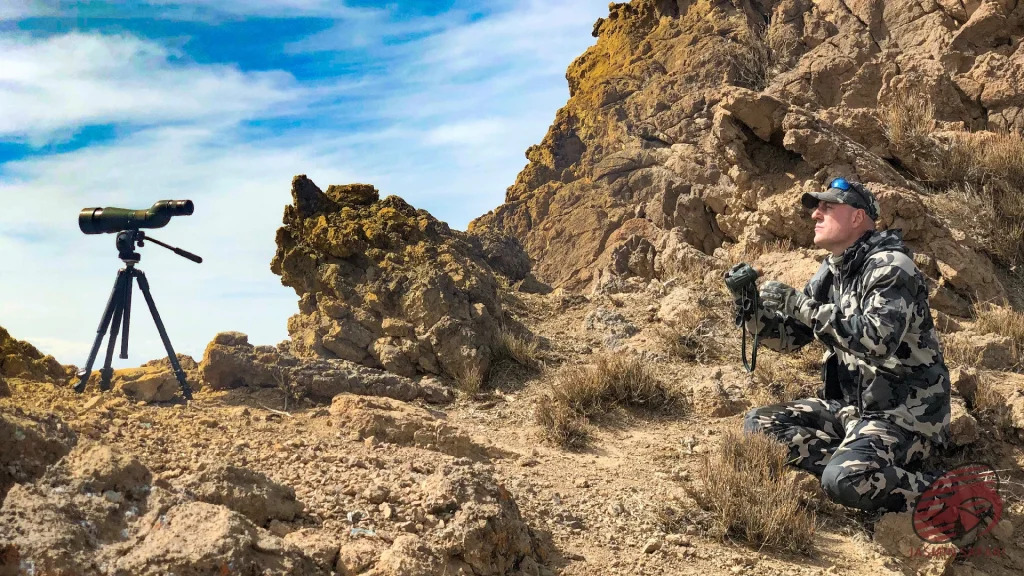 Hunter with binoculars beside a tripod spotting scope on a rocky ridge in Iran, hunt in Iran