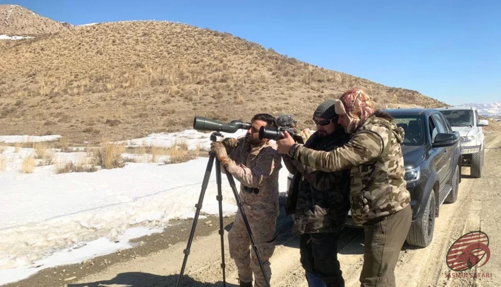 Hunters using a spotting scope beside 4×4 vehicles on a snowy roadside in Iran,